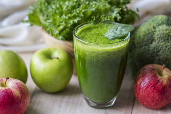 A Glass full with green juice and with some apples laying around